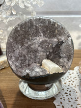 Load image into Gallery viewer, Hand-polished Brazilian amethyst geode sphere on crystal stand showing grey and lavender crystal druzy cavity with white calcite crystal inclusion
