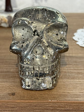 Load image into Gallery viewer, Hand-carved pyrite skull from Peru showing front view with metallic luster and black mineral inclusions on light wood surface