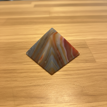 Load image into Gallery viewer, Angled view of Brazilian agate pyramid with flowing orange, grey-blue, and white banding patterns across the polished surface, 6cm base x 4.5cm height