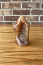 Load image into Gallery viewer, Polished carnelian agate geode tower with natural druzy cavity displaying translucent peachy-orange body and sparkling white and golden crystals on light wood surface with brick background