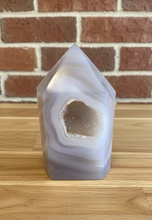 Load image into Gallery viewer, Polished white agate geode tower with heart-shaped druzy cavity displaying milky translucent body and golden-brown crystals on wood surface with brick background