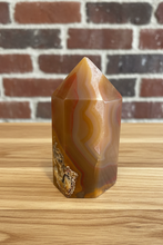 Load image into Gallery viewer, Upright view of carnelian agate tower showing natural pink and orange banding with smooth polished finish on light wood surface with brick background