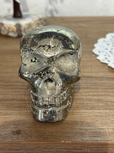 Load image into Gallery viewer, Hand-carved pyrite skull from Peru on wood shelf against brick wall, showing brilliant golden metallic luster and natural cubic crystal inclusions
