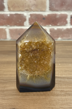 Load image into Gallery viewer, Polished citrine agate geode tower with natural golden druzy cavity displaying translucent golden-brown body and sparkling crystals on wood surface with brick background

