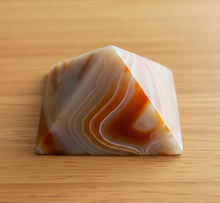 Load image into Gallery viewer, Brazilian agate pyramid with warm carnelian orange and cream banding displayed on wood table, front view showing polished finish and flowing natural patterns
