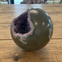 Load image into Gallery viewer, Hand-polished amethyst geode sphere on crystal stand showing side view with deep purple crystal cavity and polished olive-green exterior
