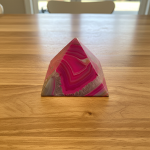 Load image into Gallery viewer, Brazilian agate pyramid with vibrant hot pink coloring and natural banding displayed on wood table, front view showing polished finish and translucent quality
