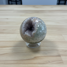 Load image into Gallery viewer, Hand-polished rainbow amethyst geode sphere on glass stand showing purple crystal cavity with natural cream and golden banding
