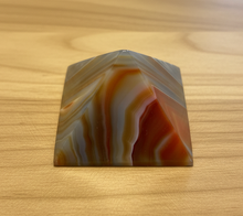 Load image into Gallery viewer, Brazilian agate pyramid with multi-colored banding in orange, grey, and blue displayed on wood table, front view showing dramatic color transitions and polished finish
